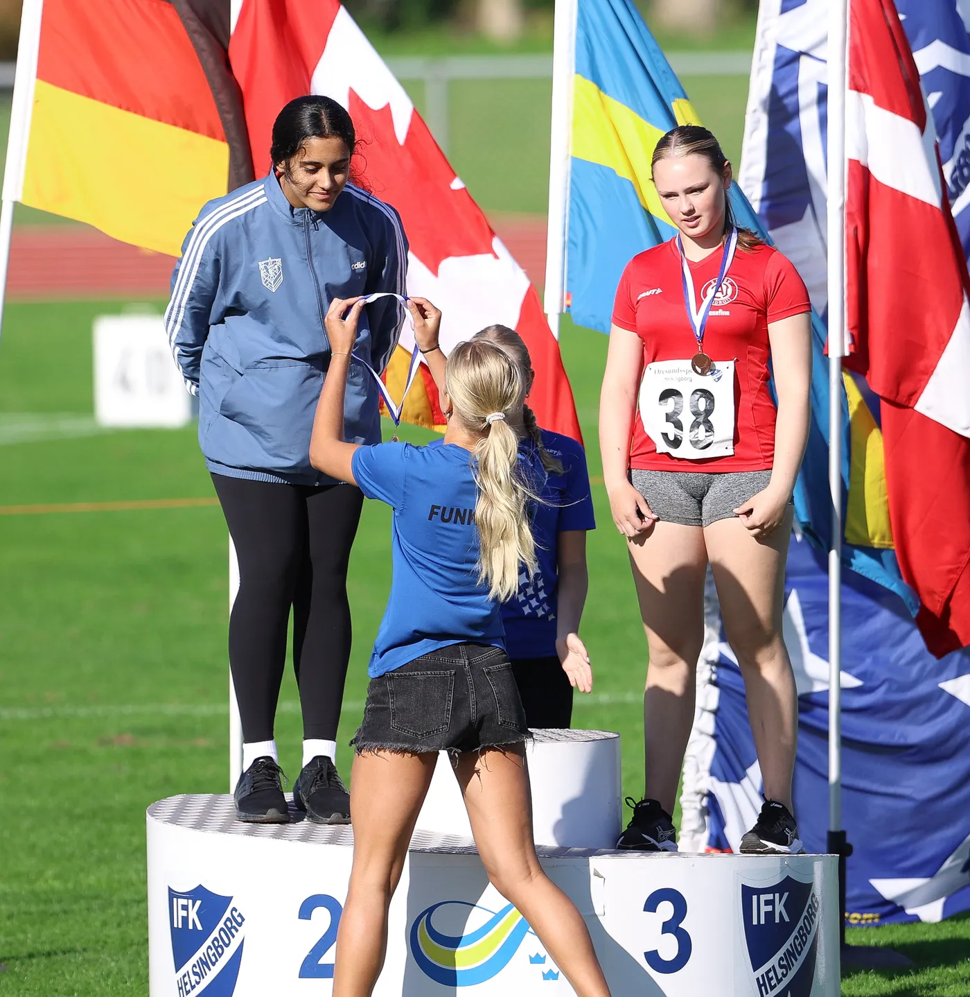 تتويج رياضي في حدث، حيث تقف شابة ترتدي سترة رياضية على منصة، باستعداد لاستلام ميدالية من فتاة ترتدي قميصًا أزرق وشورت جينز. تتواجد عدة أعلام ملونة خلفهم، تمثل دولًا مختلفة.