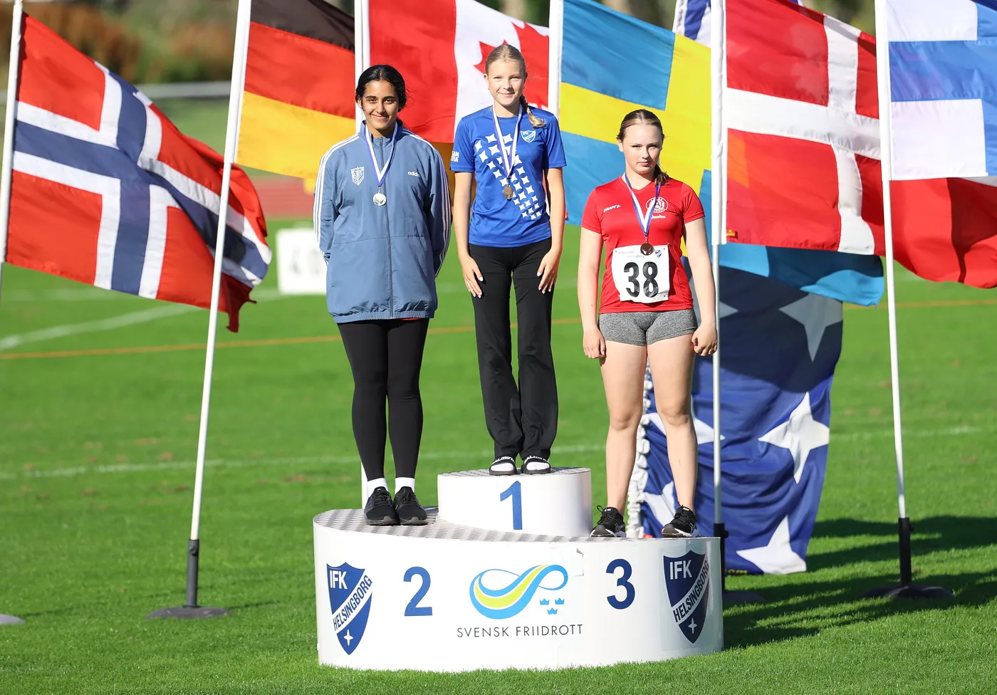 تعبّر الصورة عن ثلاث رياضيات يقفزن على منصة التتويج في حدث رياضي. الفتيات يرتدين ملابس رياضية، ويحملن ميداليات، وتقف خلفهن أعلام دول متعددة.