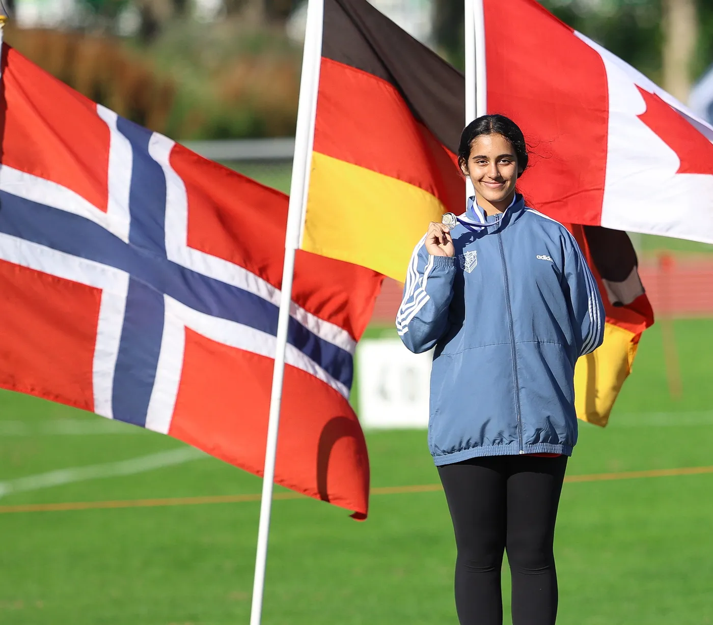 شابة ترتدي سترة رياضية زرقاء وسروالاً أسود، تحمل ميدالية ذهبية وتبتسم بينما تقف بين أعلام تتضمن أعلام النرويج وألمانيا وكندا على خلفية ملعب رياضي.