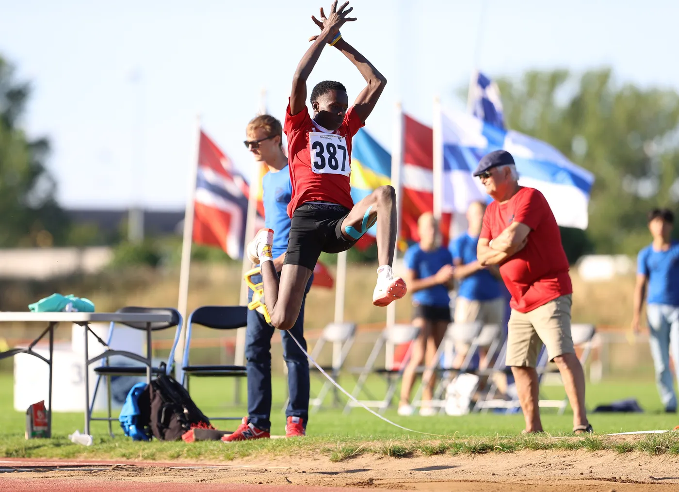 يرتفع رياضي في الهواء خلال محاولته القفز الطويل، مرتديًا قميصًا أحمر يحمل الرقم 387، بينما يشاهد مجموعة من المشجعين والمتنافسين في الخلف. تتواجد أعلام متعددة في الخلفية، مما يضيف طابعًا دوليًا للحدث.