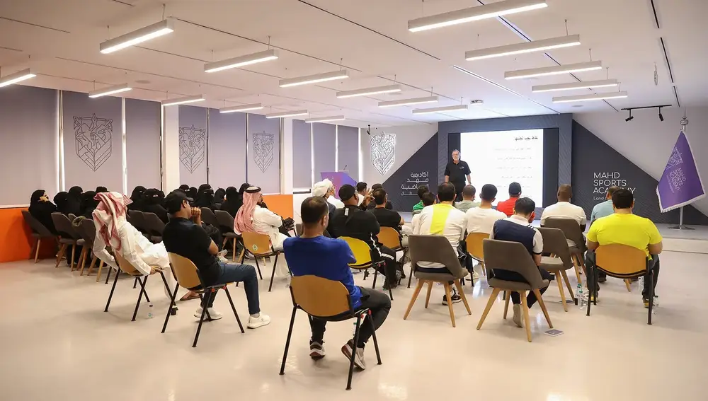 مجموعة من الأشخاص يجلسون في قاعة محاضرات، حيث يتحدث متحدث على المنصة أمام شاشة تعرض محتوى. يتواجد المشاركون في أماكنهم، بينهم رجال ونساء، ويستخدم البعض هواتفهم الذكية. يظهر في الخلفية شعار أكاديمية ماهد الرياضية.