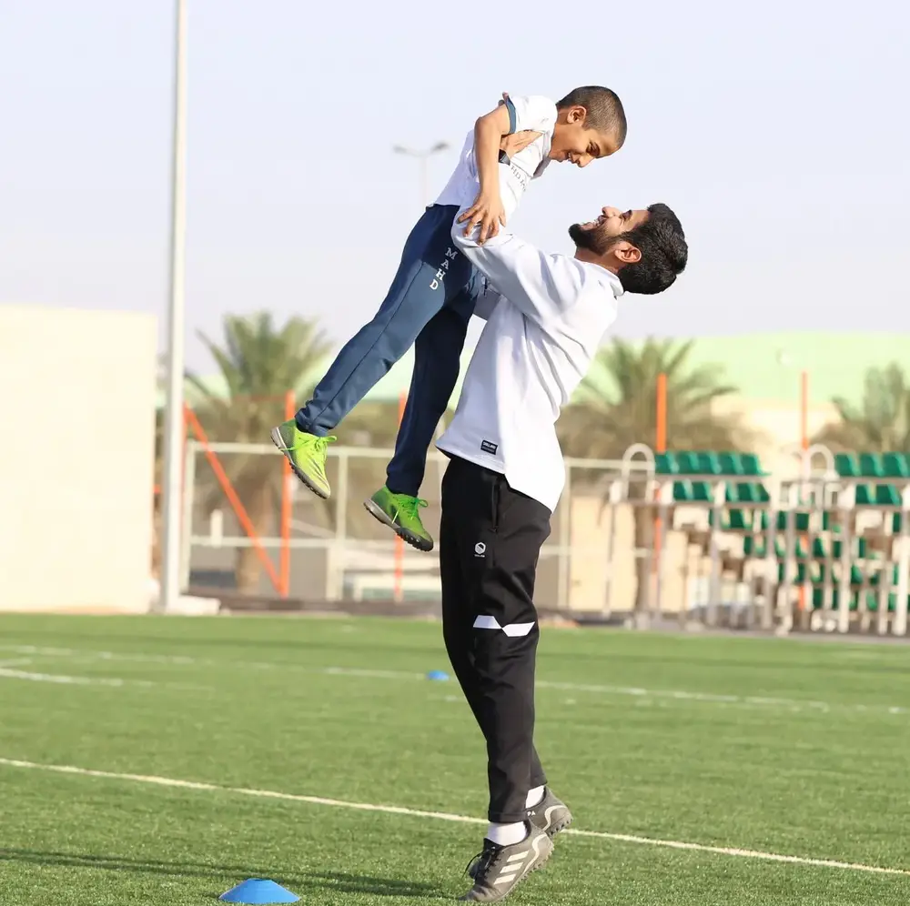 أب يحمل طفلاً صغيراً بفرح في ملعب خارجي. يبدو كلاهما سعيدين ويتبادلان الابتسامات تحت سماء زرقاء، مع وجود بعض أشجار النخيل في الخلفية.