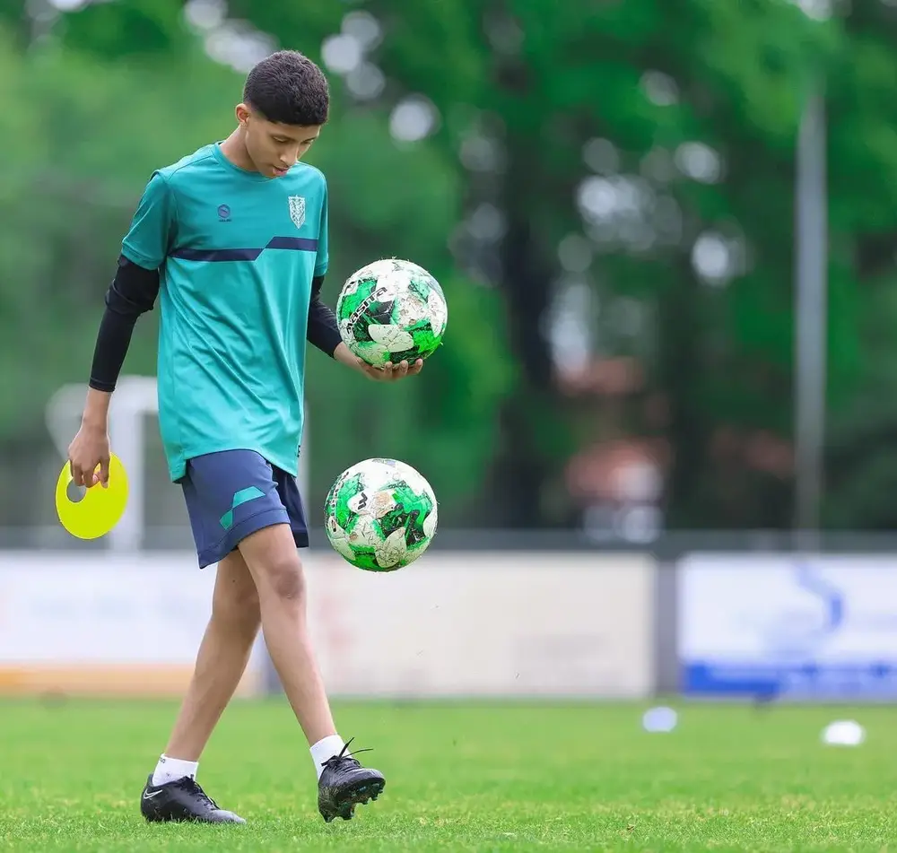 شاب يرتدي زي رياضي أزرق يقوم بالمشي على أرضية ملعب كرة القدم حاملاً كرتين خضراويتين في يديه. في اليد الأخرى، يحمل طبقاً دائرياً أصفر.