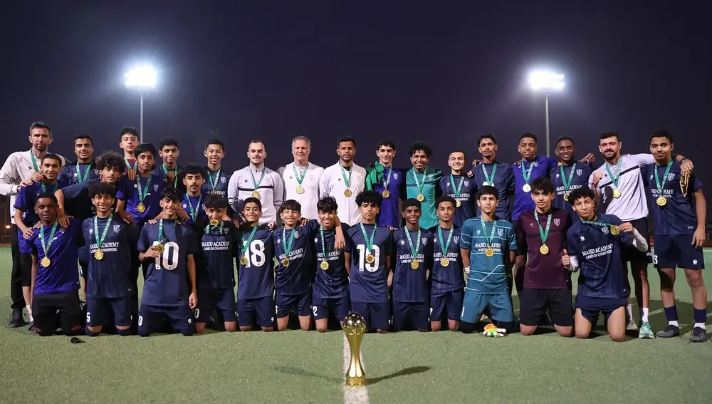 فريق لكرة القدم يتجمع بعد مباراة الفوز، مع كأس مميز في المقدمة. يرتدي اللاعبون قمصاناً زرقاء ويحملون ميداليات ذهبية، بينما يقف التدريب والجهاز الفني خلفهم تحت الأضواء.