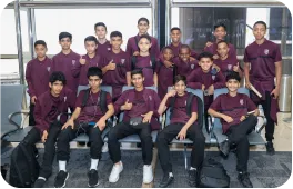 مجموعة من الشباب يجلسون في مطار، يرتدون قمصان بوردو وسراويل سوداء. يُظهر البعض إشارة الإبهام للأعلى بينما يبتسم الآخرون، مما يعكس أجواء من الحماس والترقب.