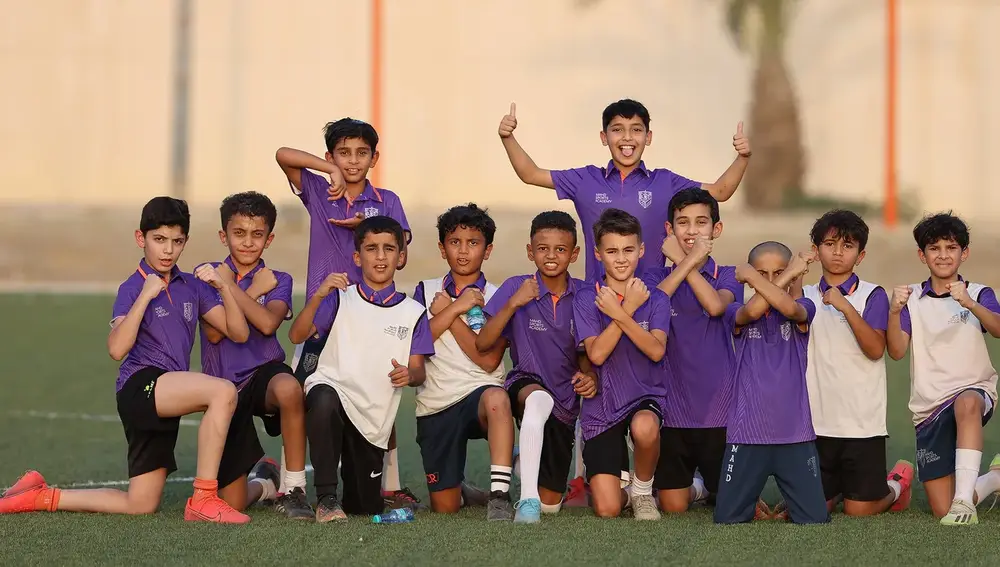 مجموعة من الأطفال الذكور في ساحة رياضية، يرتدون قمصانًا بنفسجية ويبتسمون. بعضهم يقوم بإشارات مختلفة بأيديه، بينما يقف البعض على ركبتيه، مما يدل على حماسهم ونشاطهم.