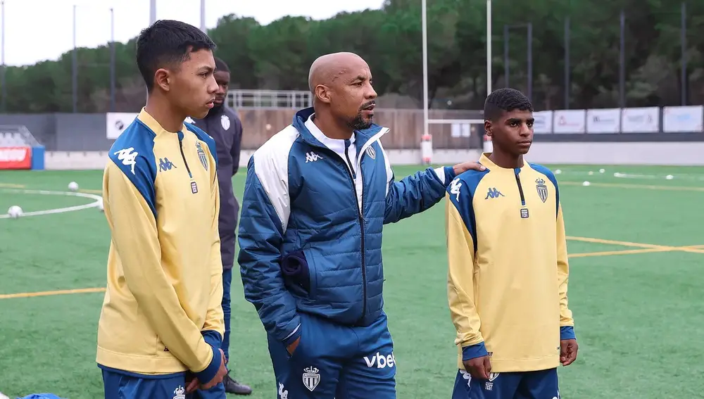 مدرب يتحدث مع لاعبين شابين على أرضية ملعب. يرتدي المدرب سترة زرقاء، بينما يرتدي اللاعبان قمصان رياضية صفراء.